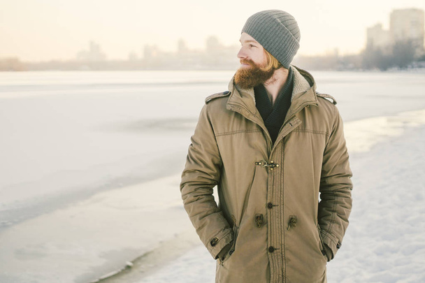 Primer plano Caucásico joven pelo rojo y barba en un sombrero y un abrigo de parque posando modelo de invierno sobre un fondo de un lago para congelar la nieve
. - Foto, imagen