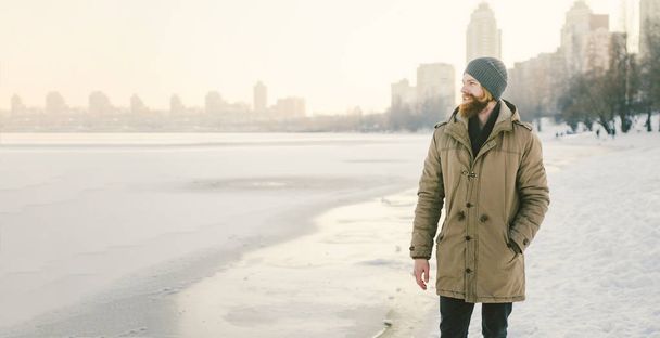 Lähikuva Valkoihoinen nuori mies punaiset hiukset ja parta hattu ja puisto takki poseeraa talvi malli taustaa vasten järven jäätyä lumi
. - Valokuva, kuva