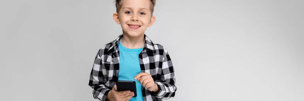 A handsome boy in a plaid shirt, blue shirt and jeans stands on a gray background. The boy is holding a phone - Фото, изображение
