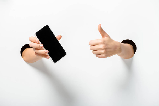 cropped image of woman holding smartphone with blank screen and showing thumb up through holes on white - Foto, imagen