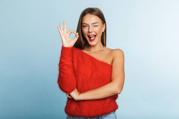 Lovely young woman wearing sweater standing isolated over blue background, showing ok gesture - 写真・画像