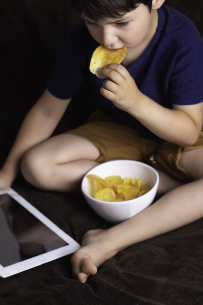 a boy holds a tablet, a gadget and eats potato chips, a concept of harmful food and dependence on gadget - Photo, Image