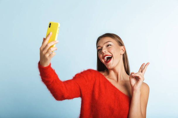 Image of a beautiful emotional excited young woman posing isolated over blue wall background take a selfie by mobile phone. - Photo, image