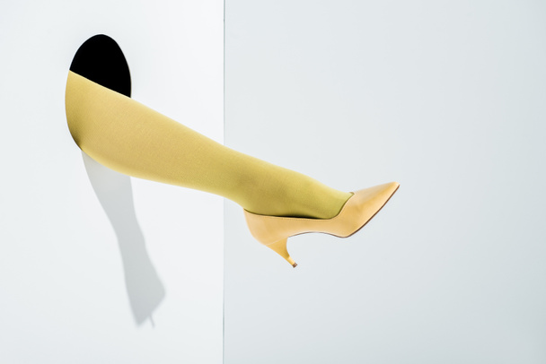 imagen recortada de niña mostrando la pierna en medias de color amarillo brillante y tacón alto en el agujero en blanco
 - Foto, Imagen