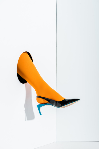 обрезанное изображение девушки, показывающей ногу в ярко-оранжевых колготках и черный высокий каблук в отверстии на белом
 - Фото, изображение