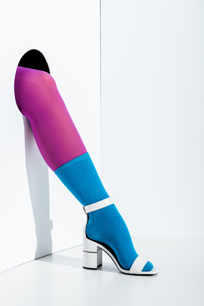 cropped image of girl showing leg in violet tights, blue sock and white high heel in hole on white - Фото, зображення