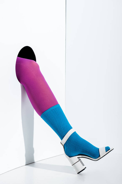 cropped image of woman showing leg in violet tights, blue sock and white high heel in hole on white - Foto, immagini