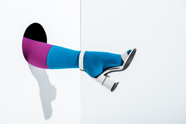cropped image of girl showing leg in trendy violet tights, blue sock and white high heel in hole on white - Foto, Imagem