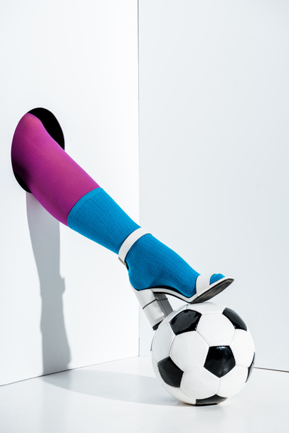 cropped image of girl putting leg in stylish violet tights, blue sock and white high heel on football ball through hole on white - Photo, Image