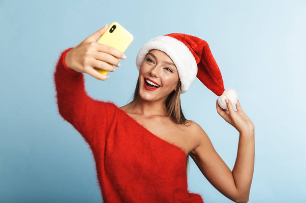 Image of a beautiful emotional excited young woman wearing christmas hat posing isolated over blue wall background take a selfie by mobile phone. - Zdjęcie, obraz
