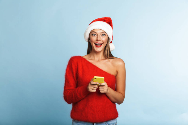 Image of a beautiful emotional excited young woman wearing christmas hat posing isolated over blue wall background using mobile phone. - Fotó, kép