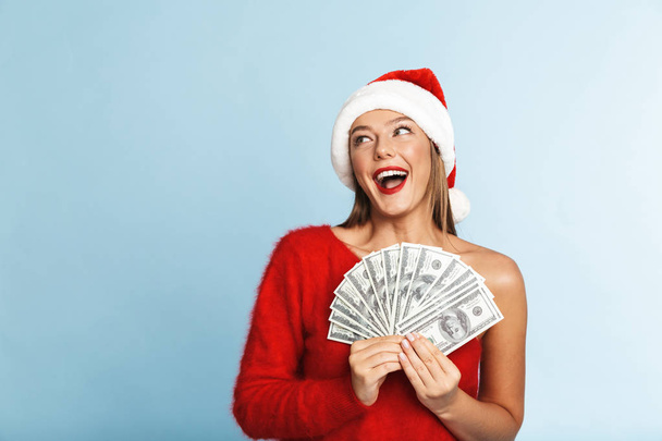 Cheerful young woman wearing santa claus hat standing isolated over blue background, showing money banknotes - 写真・画像