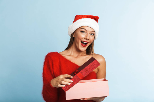Excited young woman wearing red santa claus hat standing with an open present box isolated over blue background - Fotó, kép