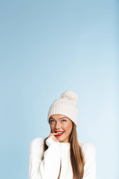 Image of a beautiful cute thinking young woman wearing winter hat posing isolated over blue wall background. - Photo, image