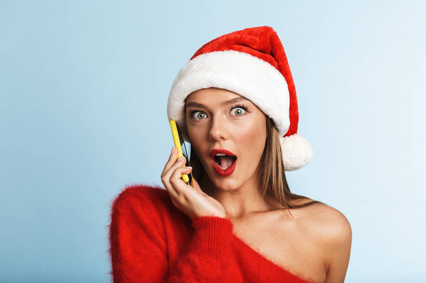 Kép egy gyönyörű érzelmi izgatott fiatal nő, fárasztó karácsonyi kalap jelentő elszigetelt keresztül, mobil telefon beszélő kék háttér. - Fotó, kép