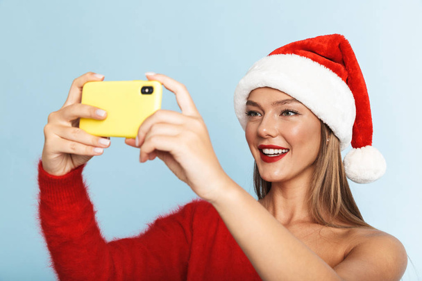 Image of a beautiful emotional excited young woman wearing christmas hat posing isolated over blue wall background take a selfie by mobile phone. - Fotó, kép