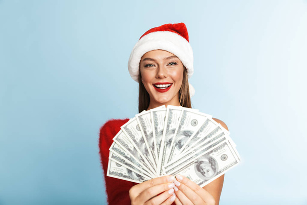 Image of a beautiful happy young woman wearing christmas hat posing isolated over blue wall background holding money. - Valokuva, kuva