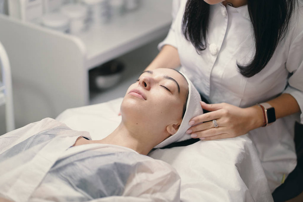 Beauty, spa and cosmetology concept. Young woman lies with closed eyes, cosmetologist making procedure for facial skin rejuvenation, facial massage. - Photo, Image