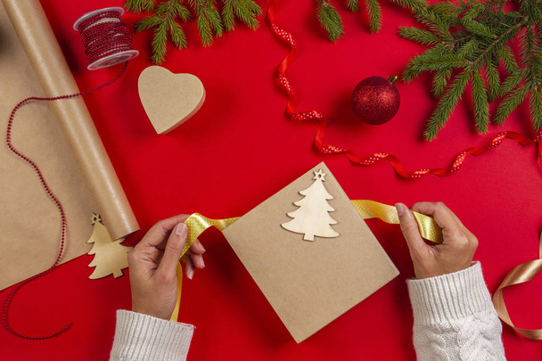 Συσκευασία δώρου Χριστουγέννων. Γυναικεία χέρια συσκευασία Χριστούγεννα παρουσιάσουν κουτί στο τραπέζι κόκκινο φόντο - Φωτογραφία, εικόνα