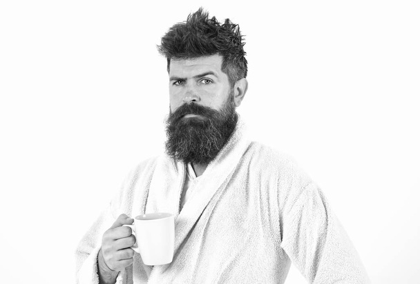 Man with beard and disheveled hair stands in bathrobe, holds mug with tea or coffee, white background. Morning rituals concept. Macho drowsy, sleepy with strict face drinks coffee in morning - Valokuva, kuva