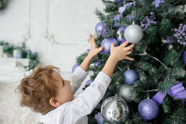 un niño rubio de 6 años decorando el árbol de Navidad con juguetes morados
 - Foto, Imagen