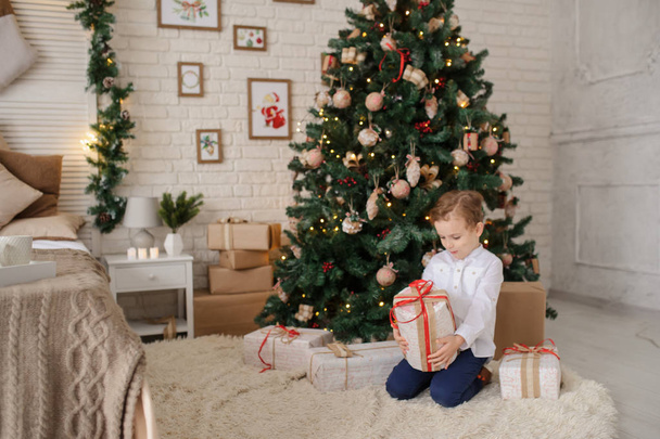ein 6-jähriger blonder Junge mit einem Geschenk in der Hand neben einem Weihnachtsbaum und einem Bett - Foto, Bild