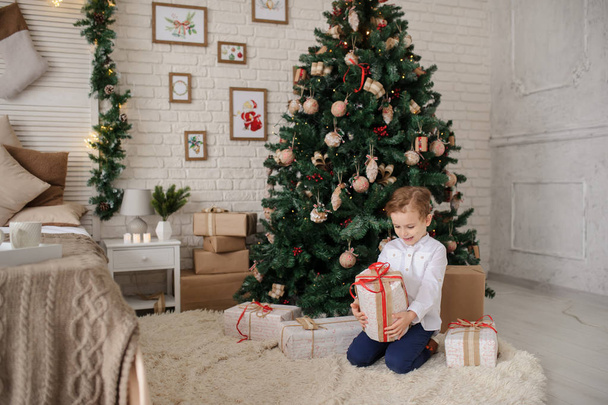 a 6-year blond boy with a present in his hand near a christmas tree and a bed - Foto, Imagen