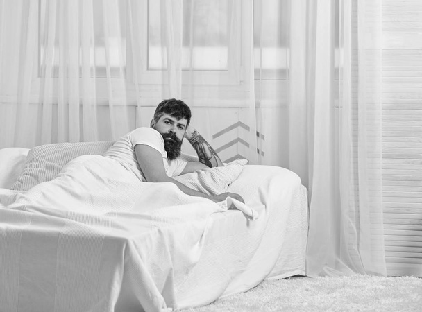 Ο άνθρωπος στο πουκάμισο τοποθέτηση στο κρεβάτι ξύπνιοι, λευκή κουρτίνα σε φόντο. Κρεβατοκάμαρα έννοια. Ο τύπος στο υπνηλία ήρεμο πρόσωπο που ξυπνάτε το πρωί. Macho με μούσι και μουστάκι, χαλαρωτικό αφού nap - Φωτογραφία, εικόνα