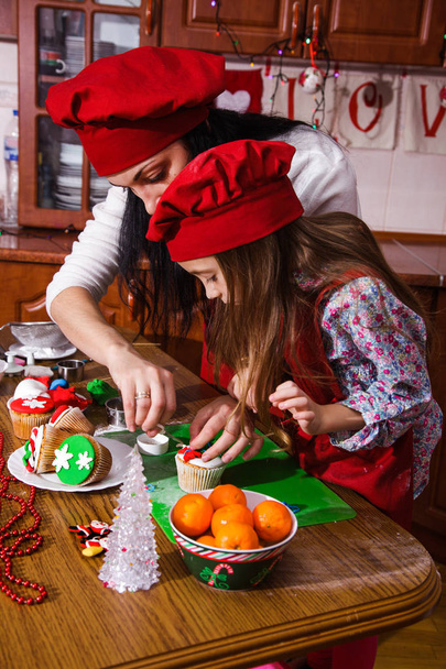 Χαμογελά ευτυχισμένος κορίτσι και μητέρα του σε ένα γιορτινό κόκκινο ποδιές μαγειρικής μπροστά από τις διακοσμήσεις Χριστουγέννων. Οικογένεια ετοιμάζει ένα επιδόρπιο για ένα νέο έτος. - Φωτογραφία, εικόνα