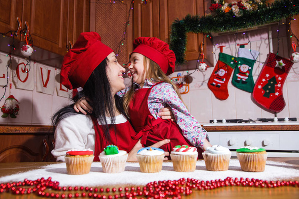 Sorrindo menina feliz e mãe em um avental vermelho festivo cozinhar na frente das decorações de Natal. Família está preparando uma sobremesa para um Ano Novo
. - Foto, Imagem