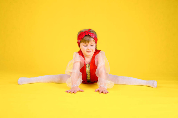 The child shows the class and sits on the splits. Yellow background. Girl with long hair in a stylized red dress - Foto, imagen