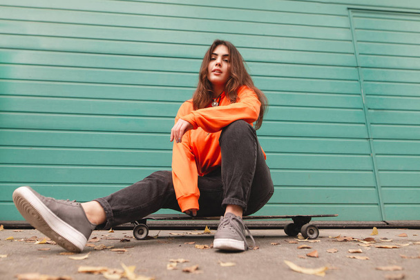 Portrait d'une jolie fille en tenue décontractée, assise sur une planche à roulettes sur un mur turquoise, regarde la caméra. Street girl assis sur un longboard
 - Photo, image