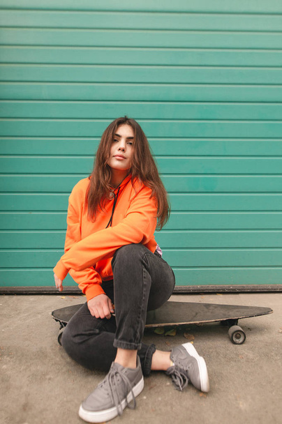 Δρόμοs κορίτσι που κάθεται σε μια longboard. Πορτρέτο του ένα ελκυστικό κορίτσι σε ρούχα, κάθεται σε ένα skateboard σε τυρκουάζ τοίχο, εξετάζει τη φωτογραφική μηχανή. - Φωτογραφία, εικόνα