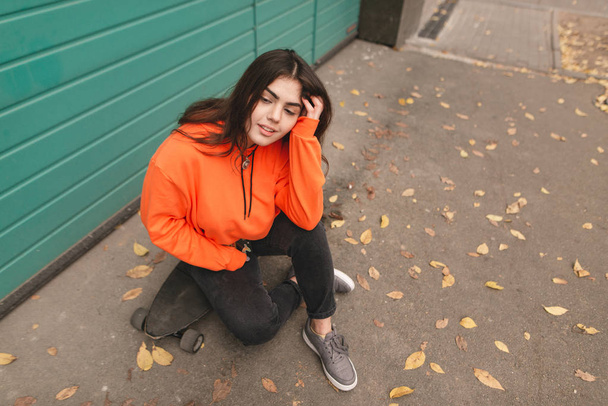 Linda menina patinadora em roupas de rua sentado na rua e sorrindo. Retrato de rua de uma menina elegante em um capuz laranja sentado em um longboard. Estilo de rua menina adolescente
 - Foto, Imagem