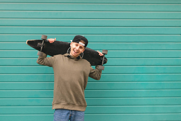 Smiling teenager in street clothes stands with a longboard in his hands against the background of a turquoise wall, looks at the camera and smiles. Copyspace - Photo, Image