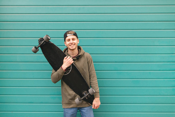 Happy patinador en ropa casual se para con un longboard en la mano contra el fondo de una pared brillante de color turquesa, mira a la cámara y sonríe. Espacio de copia
 - Foto, imagen