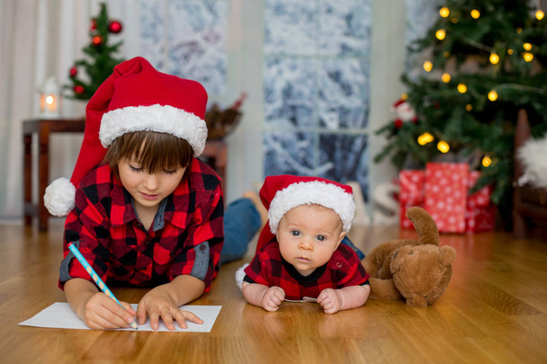 Cute newborn baby boy with Christmas hat, lying down on the floor, his older brother writing letter to Santa Claus - Photo, Image