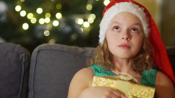 Close portrait of brown-eyed girl in santa hat with gift box in hands. Christmas celebration, new year surprise. The girl dreamily looks up - Materiał filmowy, wideo