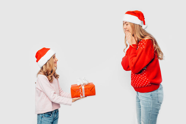 Daughter with mom in Santa hats and Christmas sweaters, daughter gives a Christmas gift to mom, on white background. - Photo, Image