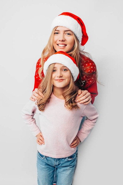 Mom and daughter are wearing Santa Claus hats, and in Christmas sweaters hug each other. On a white background. The concept of a strong family. - 写真・画像