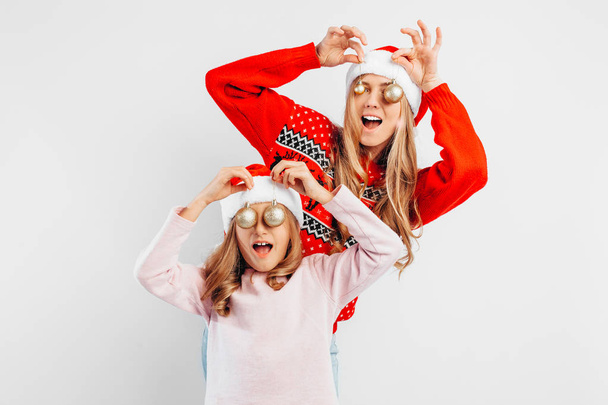 Mom and daughter are wearing Santa Claus hats, in New Year's sweaters, celebrate the New Year by playing with Christmas balls. On a white background. - Photo, image