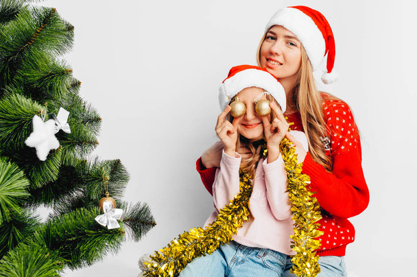 Mom and daughter are wearing Santa Claus hats, in New Year's sweaters, celebrate the New Year by playing with Christmas balls. On a white background. - Valokuva, kuva