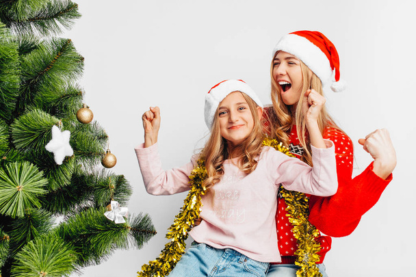 Happy mom and daughter, in Santa's hats, sitting near the Christmas tree, rejoice, showing victory gesture, on white background. - Фото, зображення