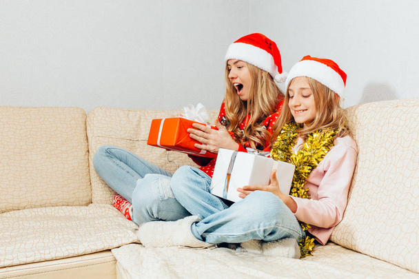 Joyful mom and daughter in Santa Claus hats are sitting on the couch watching their Christmas presents. Christmas concept - Foto, Imagen