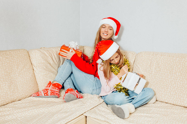 Joyful mom and daughter in Santa Claus hats are sitting on the couch watching their Christmas presents. Christmas concept - Foto, Imagen
