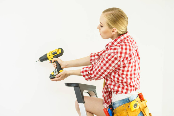 Young woman with checked shirt using drill for her work at home. Girl working at flat remodeling. Building, repair and renovation. - Foto, immagini