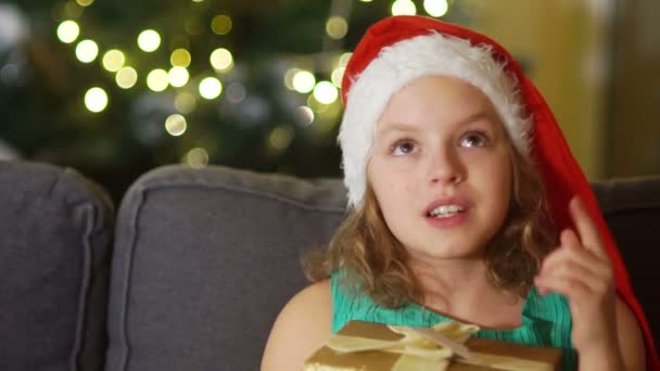 The girl makes a wish for Christmas. Close portrait of brown-eyed girl in santa hat with gift box in hands. Christmas celebration, new year surprise - Footage, Video