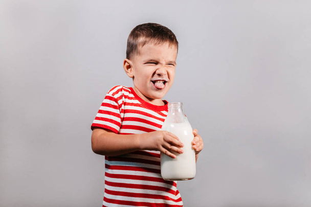 Child with bottle of milk against grey background. Portrait of young boy with milk mustache making faces isolated over gray background - studio shot. - Photo, Image