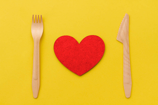 A serving of red heart with a wooden fork and wooden knife. Concept on the theme of love and restaurant on a yellow background. Fork, knife and heart. - Photo, Image