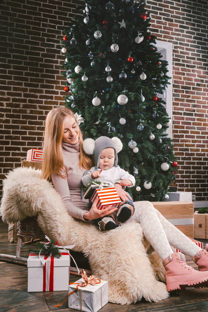 Тема різдвяні свята зимового новий рік. Молоді матері стильний кавказьких тримає її син на руках на 1 рік в смішні сорочку, сидячи на на санях і носіння хутра під дерево оформлений будинок - Фото, зображення